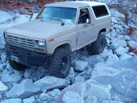 1985 Ford Bronco - A tan 1980s Ford Bronco with a white roof is navigating a rocky, off-road terrain. The vehicle has large, aggressive tires and a custom fro…