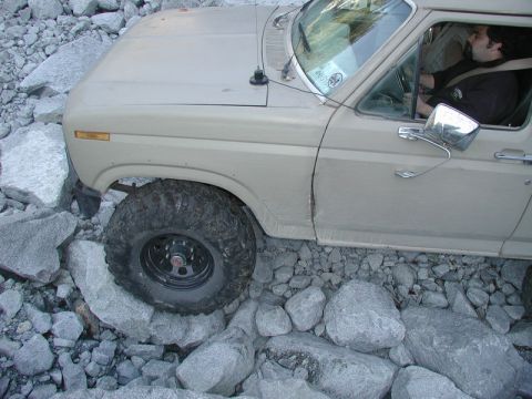 1985 Ford Bronco - A tan Ford Bronco is navigating a rocky terrain, with its front tire perched on a large boulder. The driver is visible behind the steering…