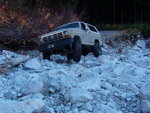 1985 Ford Bronco - A tan 1980s Ford Bronco is navigating a rocky, off-road terrain with dense trees in the background. The vehicle has large off-road tires an…