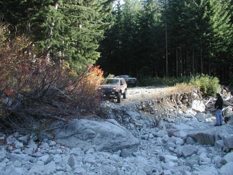 1985 Ford Bronco - A tan 1980s SUV is parked on a rocky, unpaved road next to a dense forest. Another dark SUV is visible behind it, and a person stands on th…