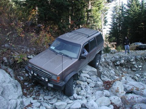 1985 Ford Bronco - A brown SUV is navigating a rocky, off-road trail with trees on either side. Another SUV is parked further up the trail.