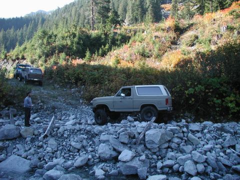 1985 Ford Bronco - A tan 1980s Ford Bronco is driving over a rocky stream bed on a dirt road, with other vehicles visible further up the road.