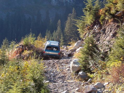 1985 Ford Bronco - A blue Ford Bronco is driving up a rocky, off-road trail surrounded by trees and boulders. The vehicle is viewed from the rear, ascending a…