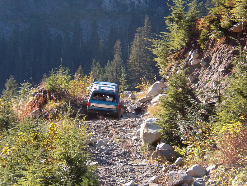 Blue 1985 Ford Bronco driving up a rocky mountain trail.