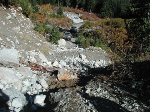 1985 Ford Bronco - Two off-road vehicles navigate a rocky, debris-strewn mountain trail with dense forest on either side. The lead vehicle is a dark-colored S…