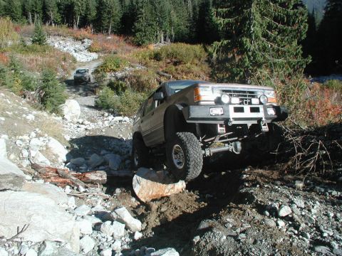 1985 Ford Bronco - A modified, lifted, tan SUV navigates a rocky, off-road trail with trees and foliage in the background. Another SUV is visible further up t…