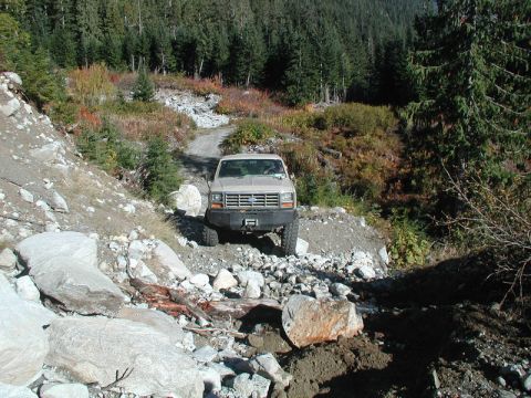 1985 Ford Bronco - A tan 1980s Ford Bronco with a custom front bumper drives on a rocky, off-road trail through a forest with autumn foliage.