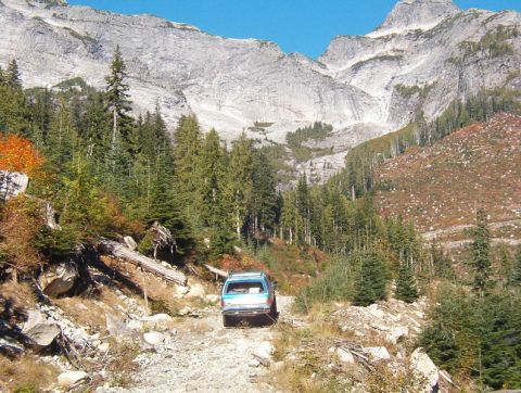 1985 Ford Bronco - A blue SUV drives up a rocky, unpaved mountain road flanked by evergreen trees and steep, rocky slopes.