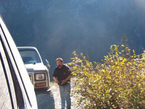 1985 Ford Bronco - A man stands next to a light-colored pickup truck on a dirt road with a wooded hillside in the background.