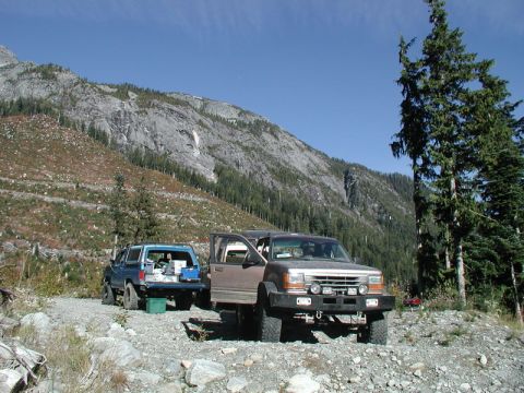 1985 Ford Bronco - Two off-road modified SUVs are parked on a gravel road in a mountainous, forested area. The vehicle in the foreground is a brown Ford Explo…