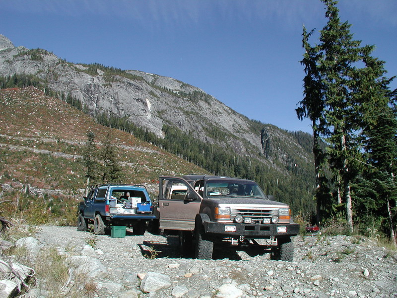 Two SUVs parked on a gravel road with mountains and trees in the background.