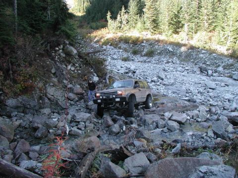 1985 Ford Bronco - A tan Ford Explorer navigates a rocky stream bed with a person standing nearby. The SUV is lifted and appears to be off-roading in a mounta…
