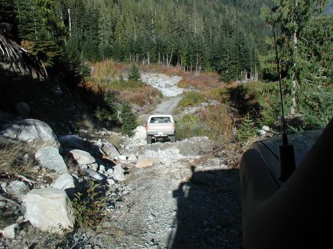 1985 Ford Bronco - A white pickup truck is driving on a rocky, unpaved road through a forest. The photo is taken from another vehicle, showing the road ahead…