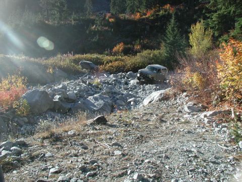 1985 Ford Bronco - A dark-colored SUV navigates a rocky, off-road terrain with autumn foliage visible on the surrounding hillsides. Another vehicle is partial…