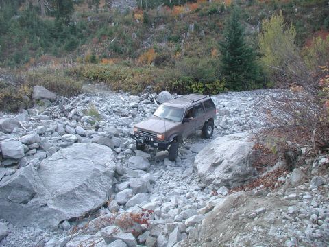 1985 Ford Bronco - A tan Ford Explorer is navigating a rocky, off-road terrain with a winch mounted on the front bumper. The vehicle is surrounded by large bo…