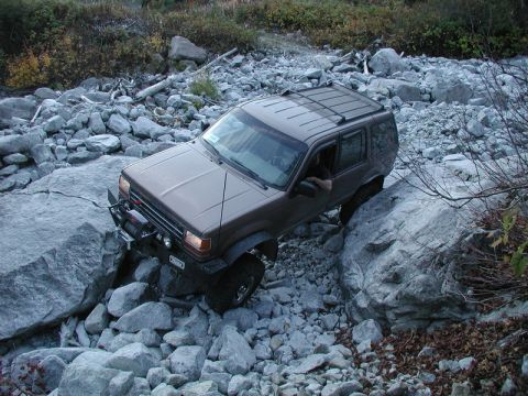 1985 Ford Bronco - A brown SUV is navigating a rocky, off-road terrain, with its front tire positioned on a large boulder. The vehicle is equipped with a winc…