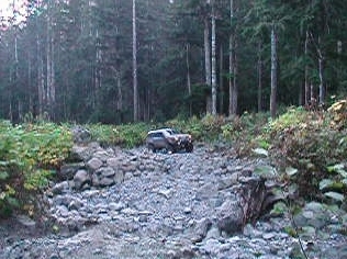 1985 Ford Bronco - A brown SUV navigates a rocky, wooded trail. The vehicle is positioned on a steep incline of loose stones and dirt.