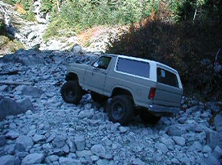 1985 Ford Bronco - A tan 1985 Ford Bronco is shown from a rear three-quarter angle, navigating a rocky, off-road terrain. The vehicle appears to be a two-door…