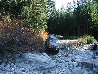 1985 Ford Bronco - A brown 1985 Ford Bronco is navigating a rocky, off-road trail next to a dense forest. Another vehicle is visible parked further up the tra…