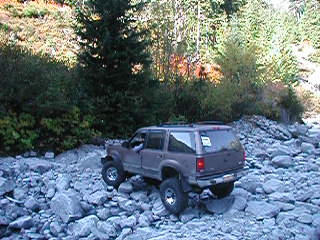 1985 Ford Bronco - A brown Ford Explorer SUV is navigating a rocky, off-road terrain with trees and foliage in the background. The vehicle is viewed from the…