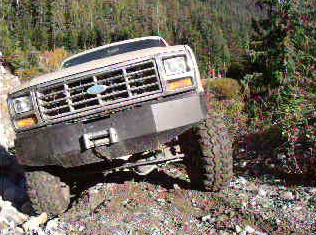 1985 Ford Bronco - A lifted 1985 Ford Bronco with a custom front bumper and winch is angled on a rocky, off-road trail. The vehicle is tilted to the right, sh…