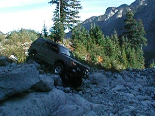 1985 Ford Bronco - A dark-colored SUV is navigating a rocky, off-road trail with steep, tree-covered mountains in the background.