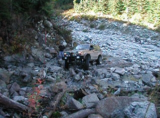 1985 Ford Bronco - A tan 1985 Ford Bronco is navigating a rocky, off-road terrain with a person standing near the driver's side door. The vehicle is equipped…