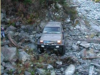 1985 Ford Bronco - A tan SUV navigates a rocky, off-road trail with dense vegetation and large boulders. The vehicle appears to be a Ford Bronco, with its fro…