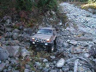 1985 Ford Bronco - A brown SUV navigates a rocky, off-road trail with dense foliage on the left. The vehicle is positioned at an angle, showing its front and…