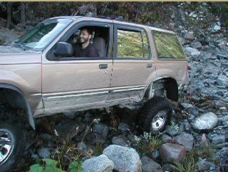 1985 Ford Bronco - A tan Ford Explorer SUV is shown navigating a rocky, off-road terrain with its front passenger side tire lifted off the ground. A man is vi…