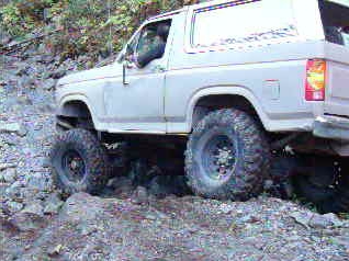 1985 Ford Bronco - A light-colored, modified 1985 Ford Bronco is shown from the side, navigating a rocky, off-road trail. The vehicle features large, aggressi…