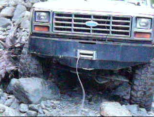 1985 Ford Bronco - The front bumper and grille of a classic Ford Bronco are visible, with a winch mounted on the bumper. The vehicle is positioned on a rocky,…