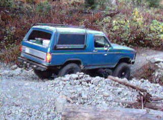 1985 Ford Bronco - A blue Ford Bronco is navigating a rocky, off-road trail. The vehicle is angled towards the right, with its rear facing slightly towards th…