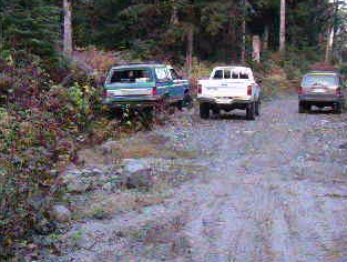 1985 Ford Bronco - A blue 1985 Ford Bronco is parked on a gravel road next to a white pickup truck and a dark SUV. The Bronco is facing away from the camera,…