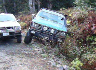 1985 Ford Bronco - A teal-colored Ford Bronco is parked on a steep, dirt incline next to a white SUV. The Bronco is angled towards the right, with its front e…