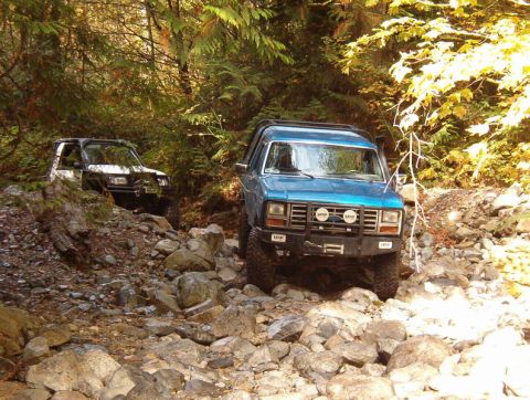 1985 Ford Bronco - A blue 1980s Ford Bronco and a white SUV navigate a rocky, wooded trail. Both vehicles are equipped for off-road driving with visible modif…