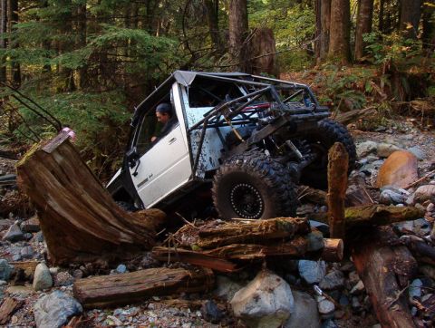 1985 Ford Bronco - A white off-road vehicle with a roll cage is navigating a rocky, wooded terrain, partially obstructed by a large fallen tree trunk. A perso…
