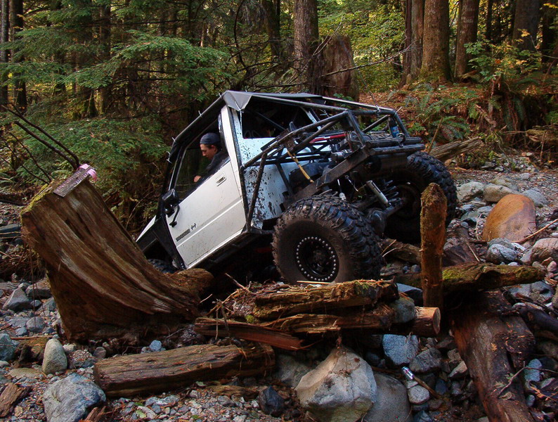 White off-road vehicle with roll cage stuck on rocks and logs in a forest.