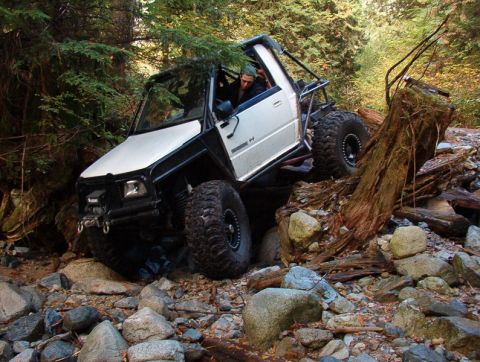 1985 Ford Bronco - A heavily modified white off-road vehicle with large tires navigates a rocky, wooded terrain with a fallen tree in the foreground.