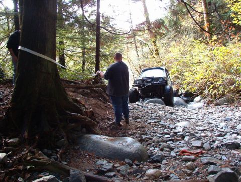 1985 Ford Bronco - A dark-colored SUV, possibly a Ford Bronco, is positioned in a rocky streambed in a forest, with two people using a winch strap attached to…