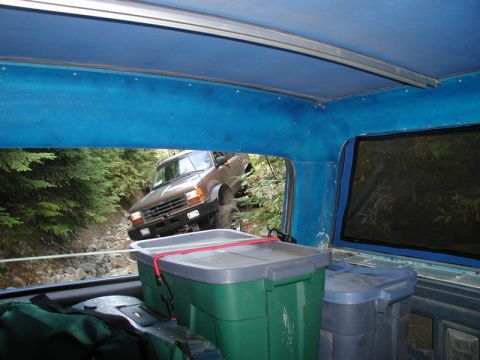 1985 Ford Bronco - View from inside a vehicle looking out at another brown SUV driving on a rocky, wooded trail. The interior has blue upholstery and two larg…