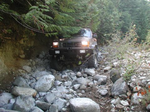 1985 Ford Bronco - A dark brown Ford Bronco is navigating a rocky, wooded trail. The vehicle is angled towards the camera, showing its front bumper and headli…