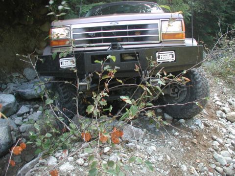 1985 Ford Bronco - The front of a lifted, tan Ford Bronco is shown navigating a rocky, wooded trail. It features a black aftermarket bumper with two "Hella" l…