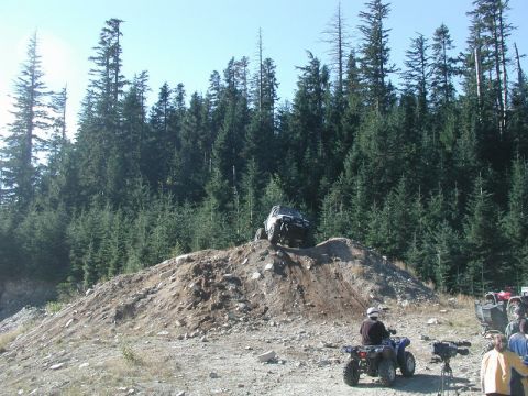 1985 Ford Bronco - A white 1985 Ford Bronco is shown climbing a dirt hill with a dense forest in the background. Several ATVs and people are gathered at the b…