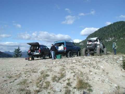 1985 Ford Bronco - Three off-road vehicles are parked on a dirt road with a mountainous, forested background. Two of the vehicles have their rear hatches open…