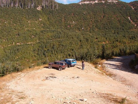 1985 Ford Bronco - Two SUVs and a van are parked on a dirt road at the edge of a dense forest, with a steep, tree-covered mountain in the background.
