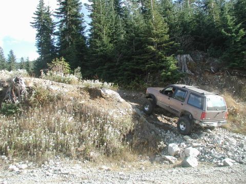 1985 Ford Bronco - A tan Ford Explorer is navigating a steep, rocky incline on a dirt trail surrounded by dense evergreen forest. The SUV has been modified wi…