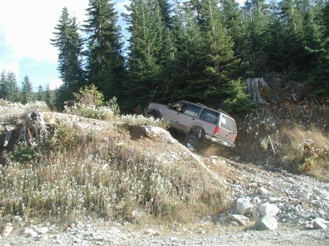 1985 Ford Bronco - A lifted, tan SUV navigates a steep, rocky, off-road trail with a dense forest in the background.