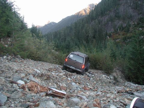 1985 Ford Bronco - A brown SUV navigates a steep, rocky, and debris-strewn mountain trail, with dense evergreen forest covering the surrounding slopes.