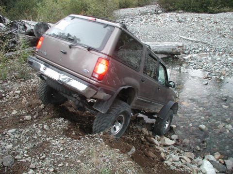 1985 Ford Bronco - A brown SUV is navigating a rocky stream bed, with its tires angled up a steep, loose dirt embankment.